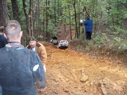 1995 Ford Bronco - Off-road vehicles navigate a challenging muddy trail through a dense forest.