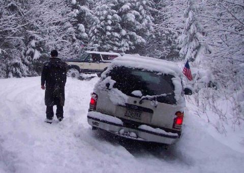 1982 Ford Bronco - Two SUVs are stuck in deep snow on a wooded road, with one vehicle displaying an American flag.