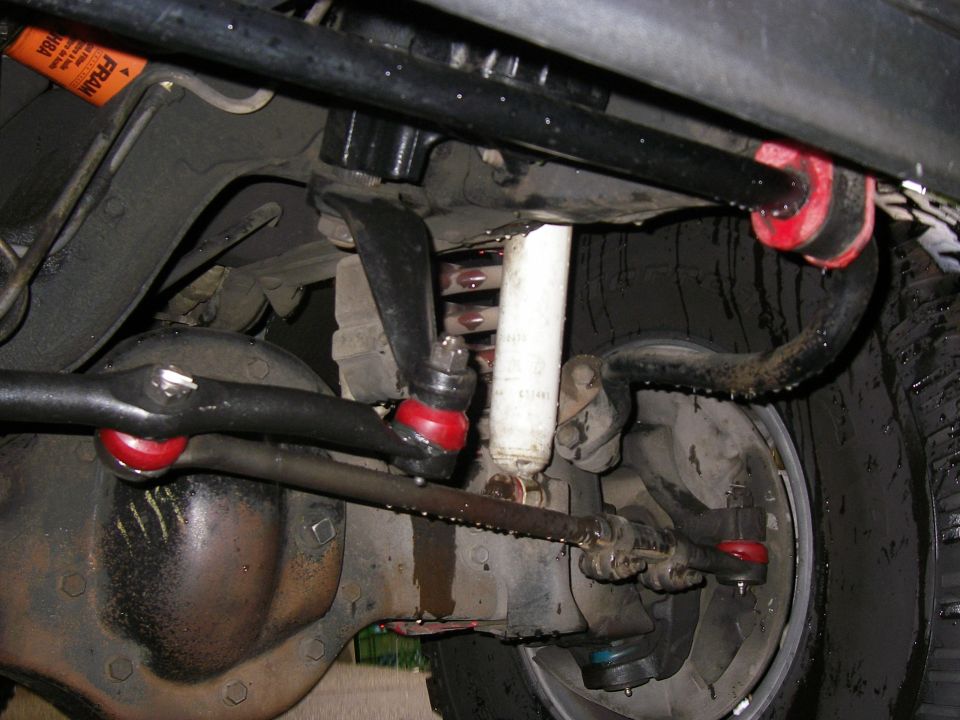Underbody view of Bronco front axle, steering linkage, white shock, coil spring, and red poly bushings.