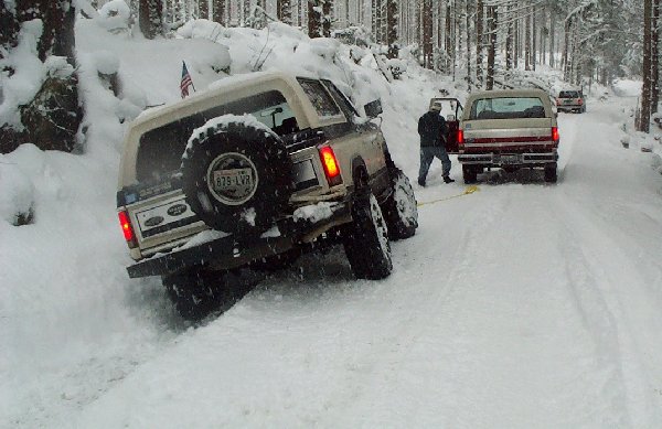 Tan and white Ford Bronco with spare tire on rear, stuck in snow, being towed by another truck