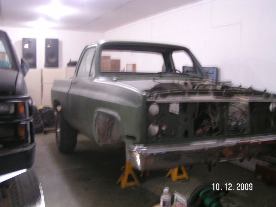 Green square-body Chevy pickup on jack stands with front end stripped in a garage