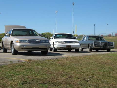 2000 Mercury Grand Marquis - Three sedans are parked in a lot on a sunny day, with one having its hood open.