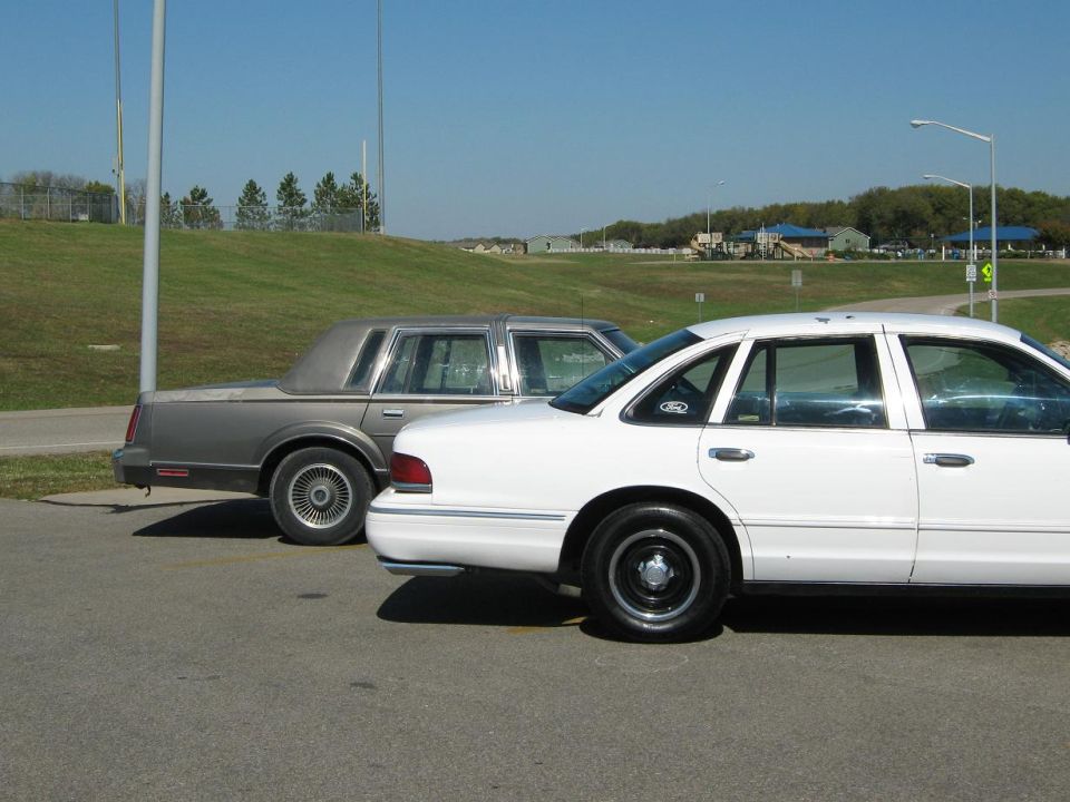White sedan and a gray sedan parked on asphalt, with a grassy hill and buildings in the background