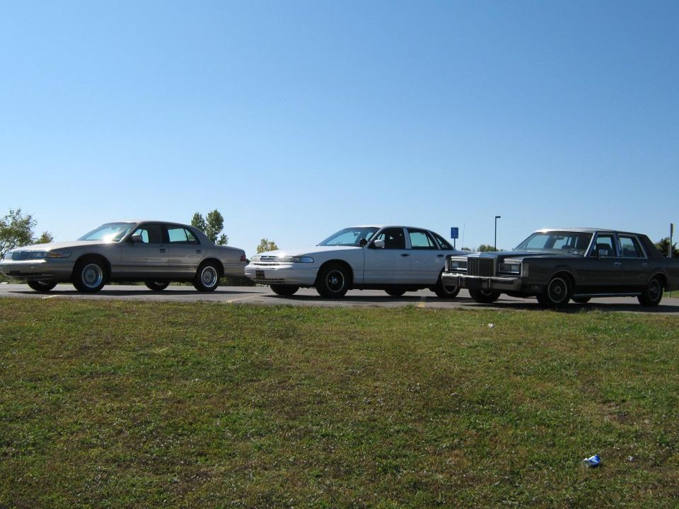 Three sedans parked side-by-side on a grassy shoulder under a clear blue sky. The leftmost car is tan, the middle is white, and the rightmost is dark gray