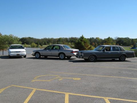2000 Mercury Grand Marquis - Three sedans are parked in a lot with a body of water and trees in the background.