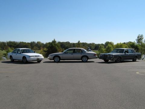 2000 Mercury Grand Marquis - A trio of sedans, including a white 1998 Mercury Grand Marquis, are parked in a lot under a clear blue sky.