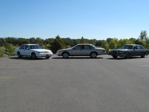 2000 Mercury Grand Marquis - Three sedans, a white Crown Victoria, a silver Mercury Grand Marquis, and a dark gray Lincoln Town Car, are parked in a lot under a clear b…