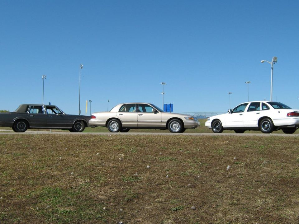 Three sedans parked on a grassy shoulder under a clear blue sky. The middle car is a tan 2000 Mercury Grand Marquis