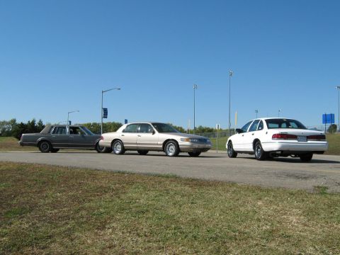 2000 Mercury Grand Marquis - A trio of sedans, including a white 2000 Mercury Grand Marquis, are lined up on the side of a road under a clear blue sky.