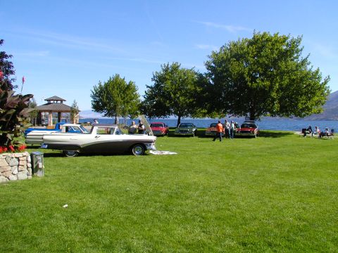 1967 Ford Ranchero - A gathering of classic vehicles is displayed on a grassy area next to a body of water under a clear blue sky.
