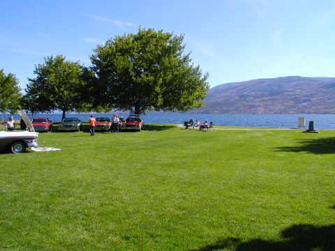 1967 Ford Ranchero - Several classic cars are gathered on a grassy area next to a large body of water under a clear blue sky.