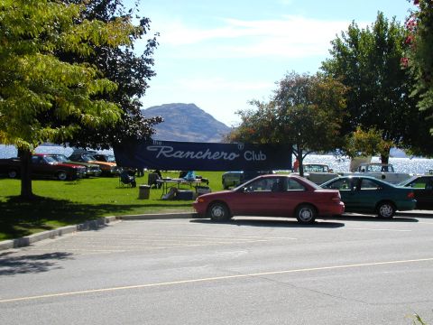 1967 Ford Ranchero - A red coupe is parked in front of a banner for The Ranchero Club, with several other vehicles on display in the background.