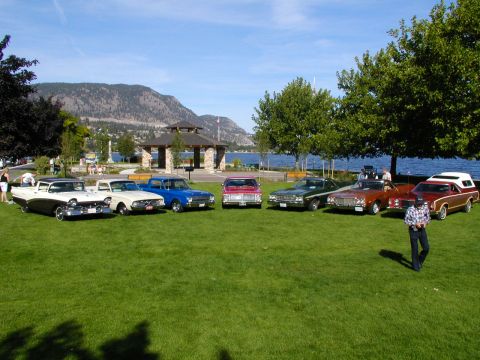 1967 Ford Ranchero - A collection of vintage vehicles, including a 1967 Ford Ranchero, are displayed on a grassy area near a scenic lake and park pavilion.