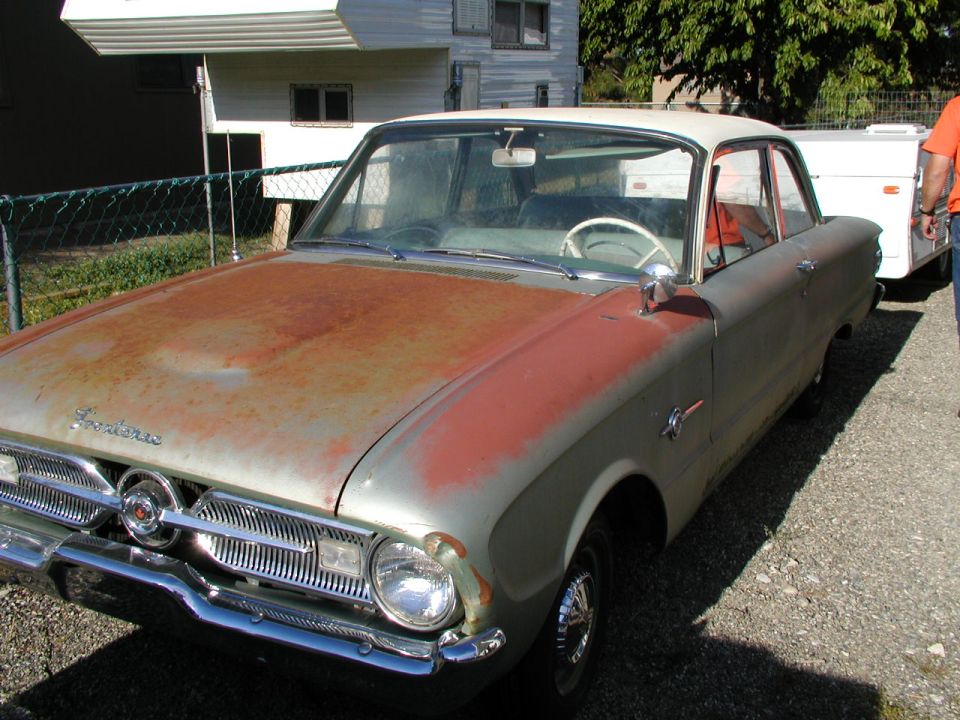 Faded green early-1960s Ford Falcon two-door with rusty hood on gravel lot