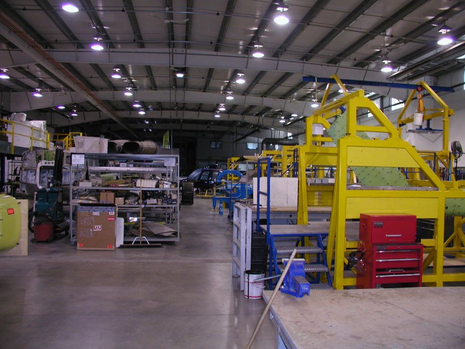 Factory interior with yellow assembly line machinery, shelves of parts, and a dark vehicle in the background