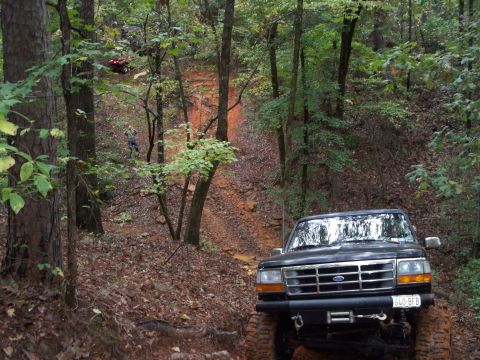 1995 Ford Bronco - A black 1995 Ford Bronco navigates a steep, muddy trail through a dense forest, with another vehicle visible further up the incline.