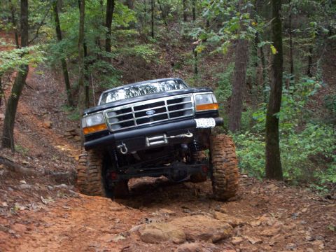1995 Ford Bronco - Front view of a lifted dark Ford Bronco descending a rocky red-clay hill, its solid front axle flexed with one mud tire drooped into a hole…