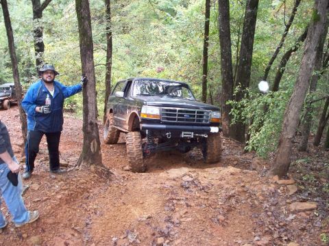 1995 Ford Bronco - A lifted Ford Bronco navigates a muddy, wooded trail with a man and another vehicle nearby.