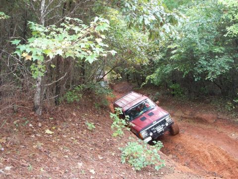 1995 Ford Bronco - A red Jeep Cherokee with a roof rack and hood louvers works down a steep off-camber red-clay descent in the woods, leaning hard toward the…