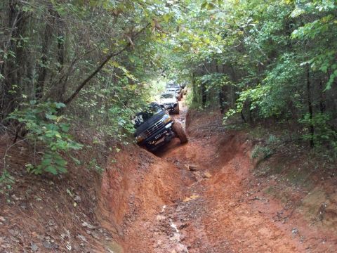 1995 Ford Bronco - A dark green lifted Ford truck with a winch bumper, roof-mounted light bar and mud tires crawls down a deeply rutted red-clay trail, body l…