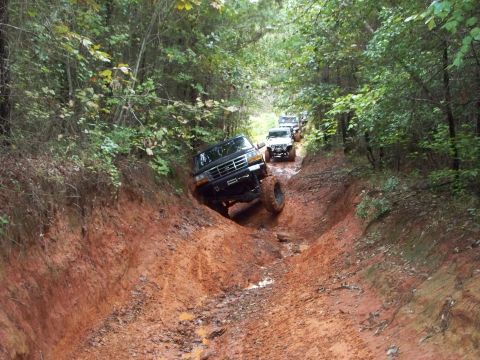 1995 Ford Bronco - Several off-road vehicles descend a challenging, muddy trail surrounded by dense forest.