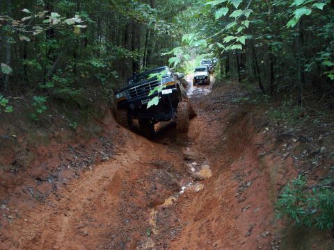 1995 Ford Bronco - A convoy of SUVs, including a 1990s Ford Bronco, tackles a challenging muddy path through a wooded area.