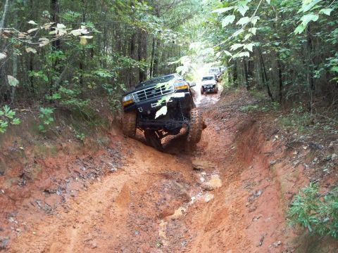 1995 Ford Bronco - Front view of a dark green lifted Ford truck nosing down a steep, deeply eroded red-clay rut, front tires crossed up with one wheel drooped…