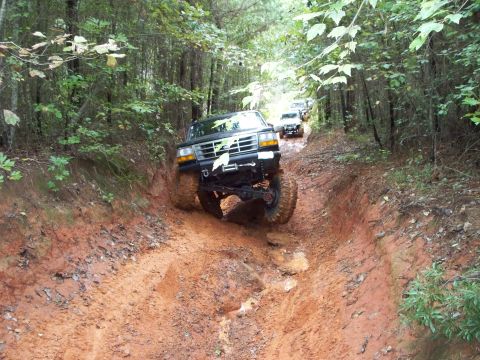 1995 Ford Bronco - A lifted black Ford SUV on mud-terrain tires drops into a deep red-clay washout on a wooded trail, front end angled down with a winch and t…