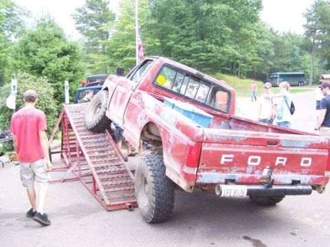 1989 Ford F-150 - Old red Ford pickup with FORD tailgate climbing a steel ramp obstacle while spectators watch at an outdoor event.