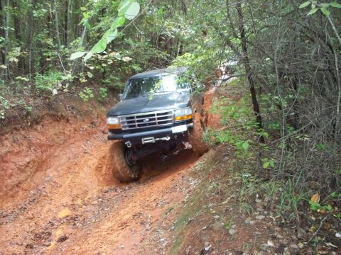 1995 Ford Bronco - A dark green lifted Ford Bronco crawling down an off-camber red-clay trail rut in the woods, front wheels flexed and body leaning hard to o…