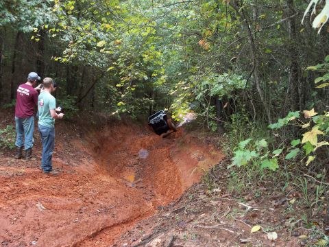 1995 Ford Bronco - A dark 4x4 climbs out of a deeply rutted red-clay trench on a wooded off-road trail, front end tipped up at a steep angle. Two spectators i…