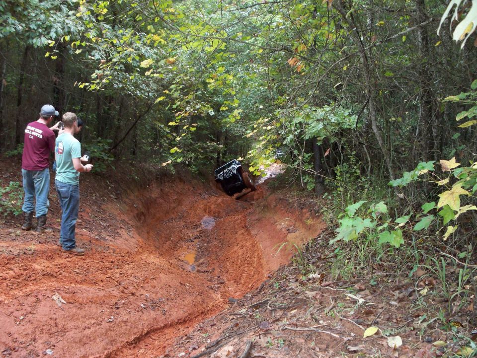 Dark 4x4 climbing out of a red-clay rut on a wooded trail as two men watch