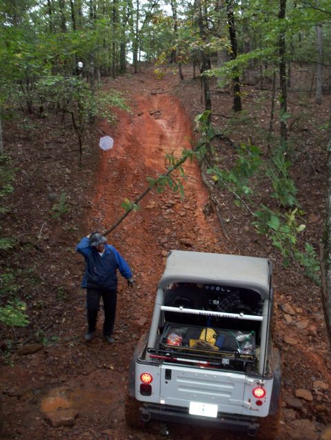 1995 Ford Bronco - A Jeep navigates a steep, muddy incline on a forest trail, with a person observing the ascent.