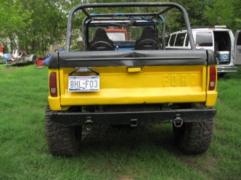 1973 Ford Bronco - Rear view of a yellow Ford Bronco with a black roll cage and bumper, parked on grass with other vehicles nearby.