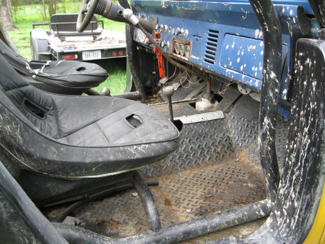 Interior of a blue Ford Bronco with aftermarket seats and roll cage. A trailer is visible in the background