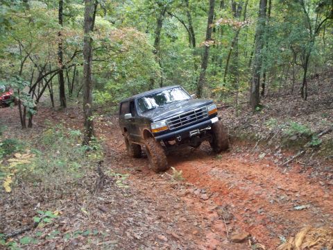1995 Ford Bronco - Three-quarter front view of a mud-splattered dark green lifted Ford Bronco climbing an off-camber red-clay bank in the woods, articulating…