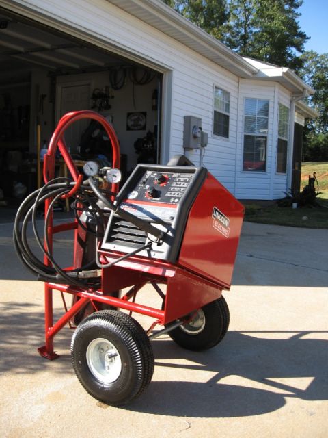 1995 Ford F-150 - A red welding cart with a Lincoln Electric welder is parked on a concrete driveway in front of a garage.