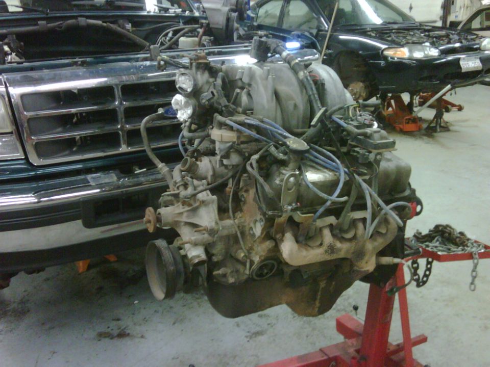 Engine on a red stand in front of a dark green vehicle with hood open