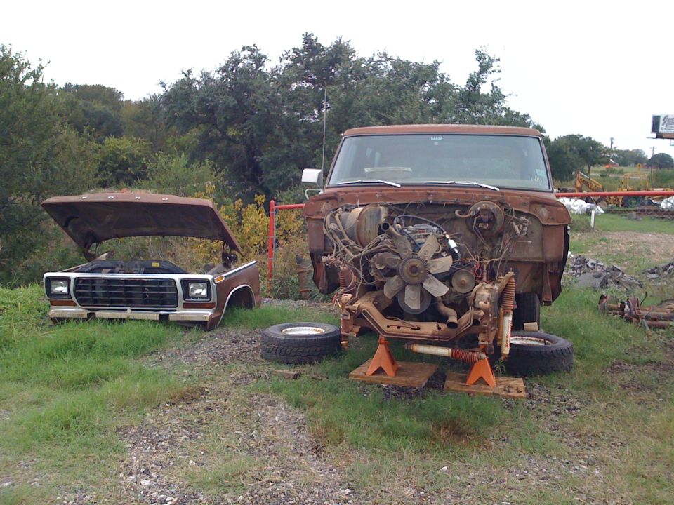 Two rusty Ford trucks, one with hood open and front clip removed, the other with hood up