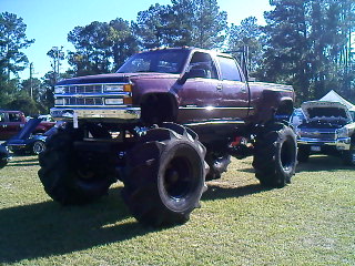 1990 Ford Bronco - A heavily modified pickup truck with massive tires and lifted suspension is displayed outdoors, with other vehicles visible in the backgrou…