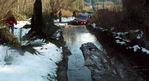 1982 Ford Bronco - Multiple vehicles navigate a challenging muddy and watery trail with snow on the sides.