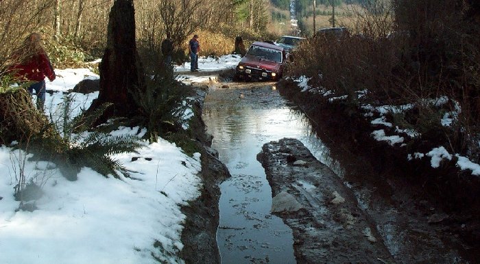 Red off-road vehicle with auxiliary lights stuck in a muddy water-filled rut, with snow on the banks