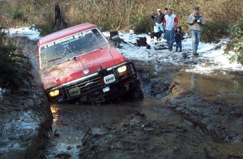 1982 Ford Bronco - A red off-road vehicle navigates a challenging muddy trail with snow patches on the ground.
