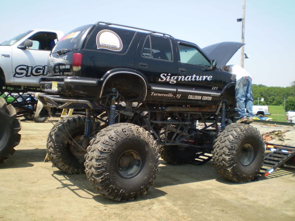 Black monster truck with large tires, lifted suspension, and "Signature Collision Center" text on the side. A person is working on the engine under the open hood. Another truck is partially visible to the left