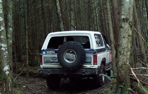 Rear view of a white and blue Ford Bronco with a spare tire mounted on the back, driving on a dirt path through a forest