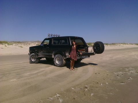 1993 Ford Bronco - A woman stands beside a black Ford Bronco parked on a sandy beach under a clear blue sky.