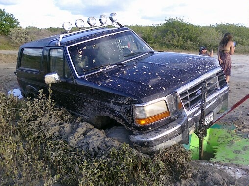 Black 1993 Ford Bronco stuck in mud with a Hi-Lift jack trying to free it. A woman stands nearby