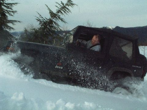 1982 Ford Bronco - A Jeep Wrangler navigates a snowy terrain, with snow spraying up around the vehicle.