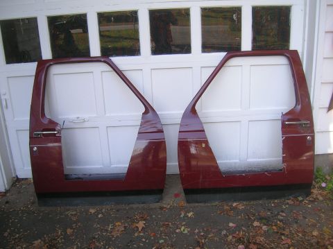 1996 Ford Bronco - A matched pair of dark red two-tone truck doors stripped of glass, interior panels and window regulators, leaning against a white garage do…