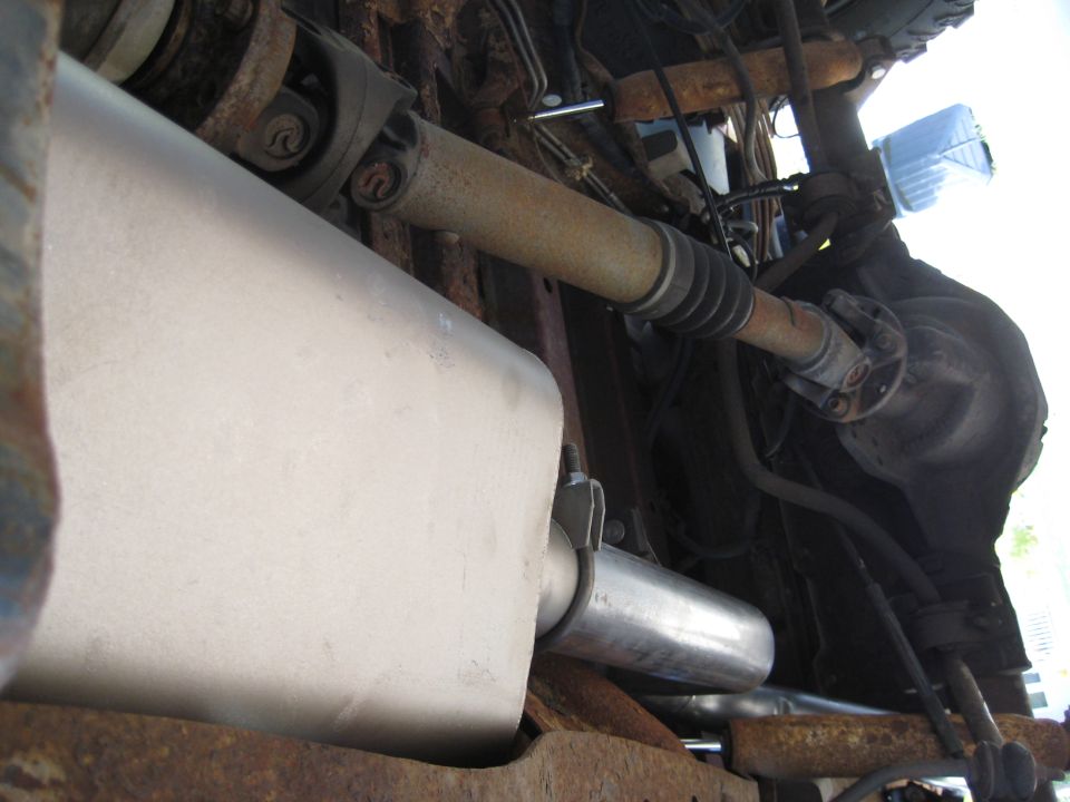 Undercarriage of a vehicle showing a muffler, driveshaft, and differential. The metal components are rusty